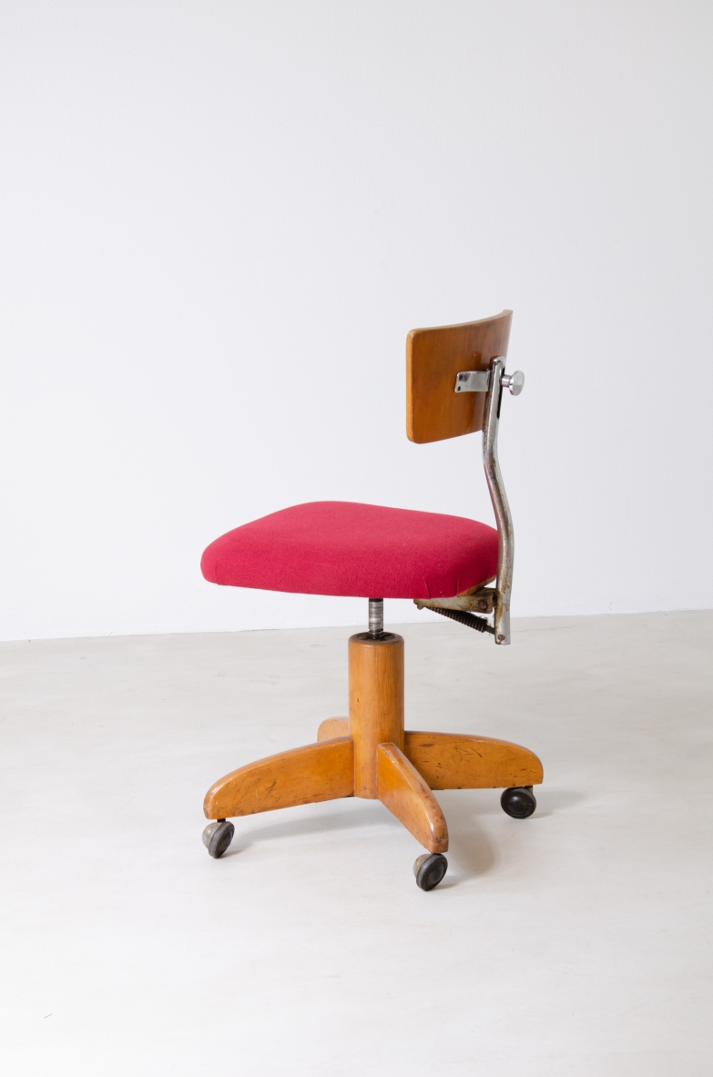 Side view of a Italian desk chair with red upholstered seat, curved wooden backrest supported by a metal frame and four-spoke wooden base with casters.