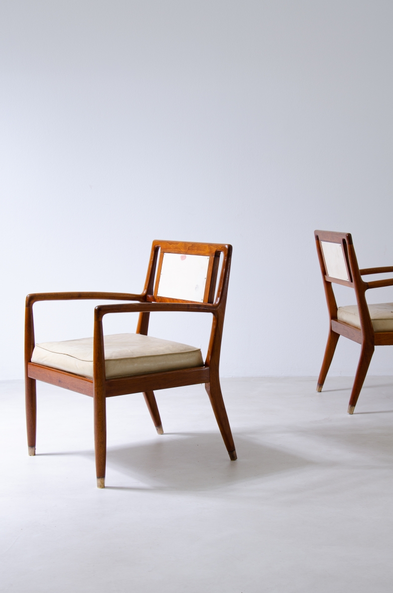 Angled view of two 1950s Italian walnut armchairs with shaped wooden armrests, light fabric-upholstered seats and backrests and brass feet.