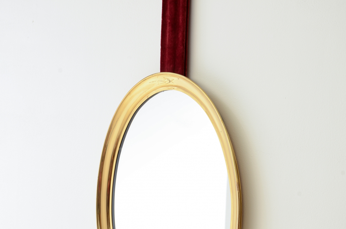 Specchiera in ottone sagomato appesa a un nastro in velluto originale e fissata a parete mediante un disco in ottone.  Manifattura italiana anni 50/60ca.