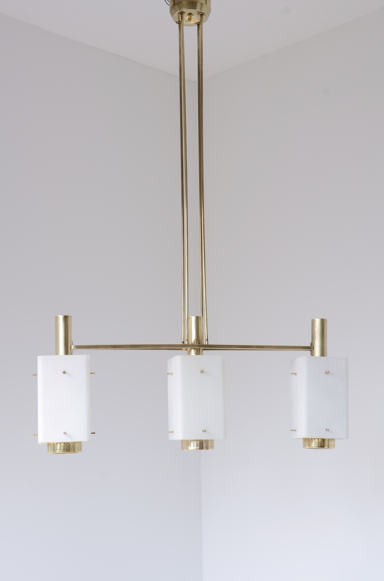 Bruno Gatta three-light chandelier for Stilnovo Milano,featuring a geometric brass frame, central suspension rod and three cylindrical white opaline glass diffusers with brass details, front view.