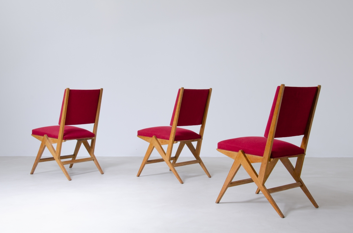Group view of three French chairs in light oak, with crossed wooden legs and red cotton-upholstered seats and backrests.