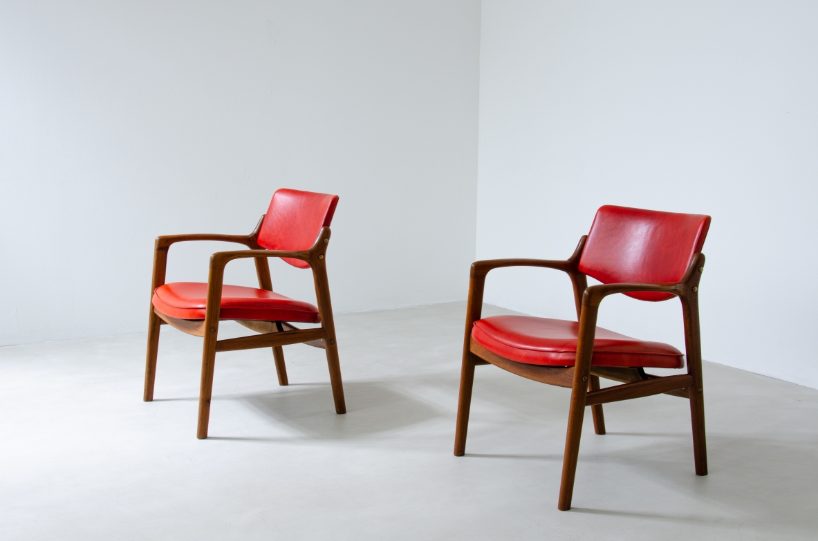 Angled view of two 1960s Danish teak armchairs with curved wooden armrests, red leather-upholstered seats and backrests, and tapered legs.