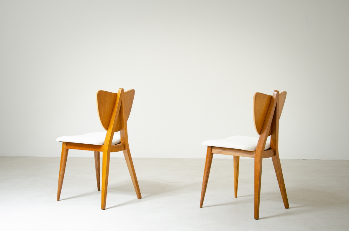 Side view of two 1950s French chairs in light oak, with heart-shaped wooden backrests, white upholstered seats and splayed legs.