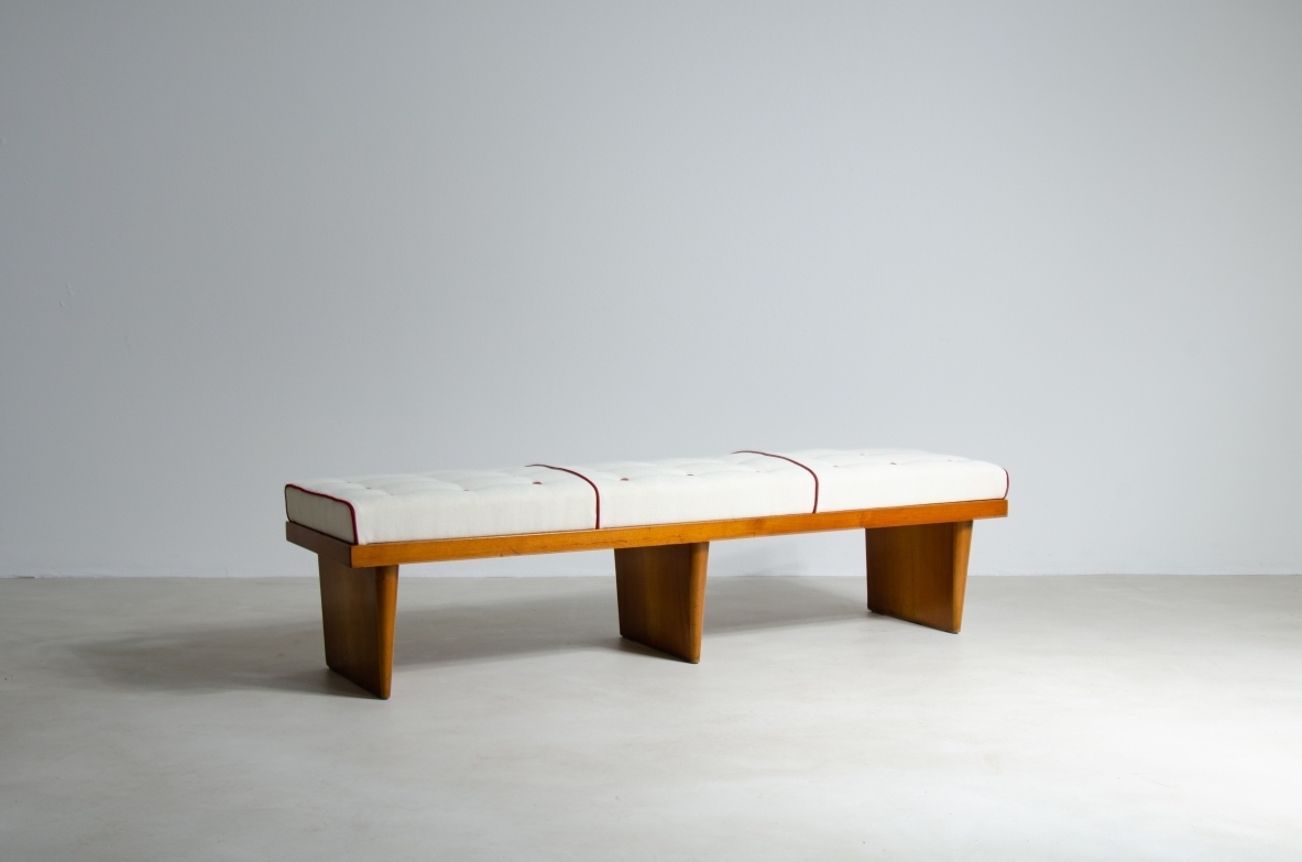 Angled view of a bench attributed to Gio Ponti, with a rectangular seat upholstered in light-colored fabric and a wooden frame with three supports.