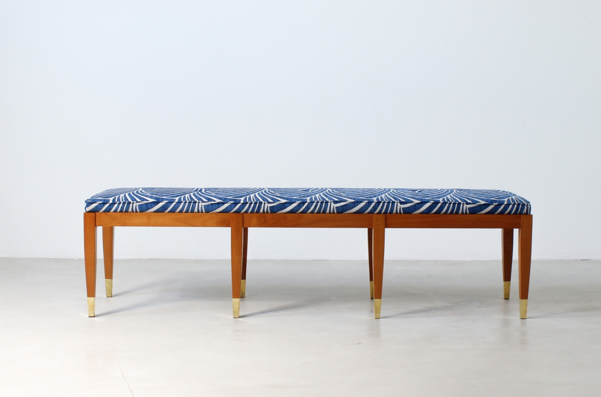 Front view of a long cherrywood bench with a blue patterned upholstered seat, six tapered wooden legs and brass feet.