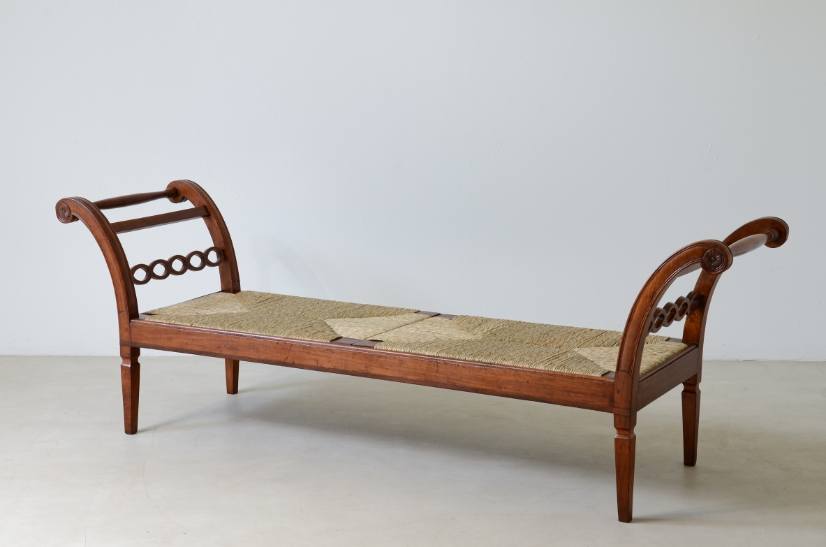 Angled view of a long 19th-century Tuscan cherrywood bench with curved ends, decorative chain motifs on the sides and a woven straw seat with a light-colored cushion.