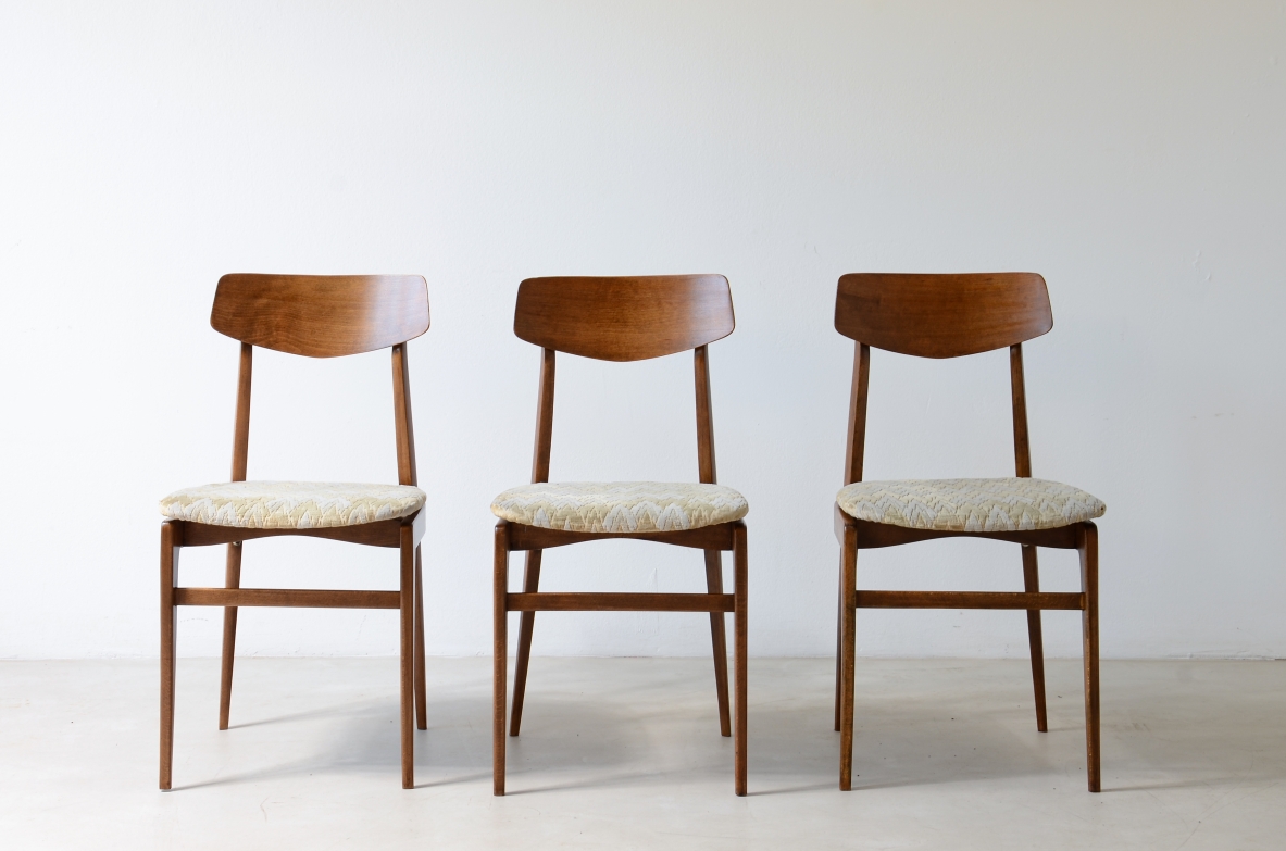 Front view of three 1960s teak chairs with open backs, curved wooden backrests and light-colored upholstered seats.