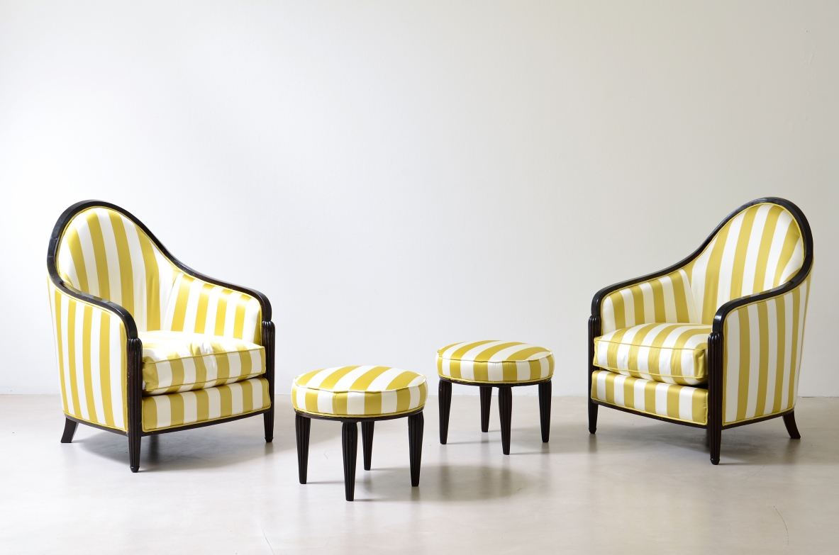 Front view of two French Art Deco armchairs with curved wraparound backs, dark wooden trim and yellow and white striped upholstery, accompanied by two matching small stools.