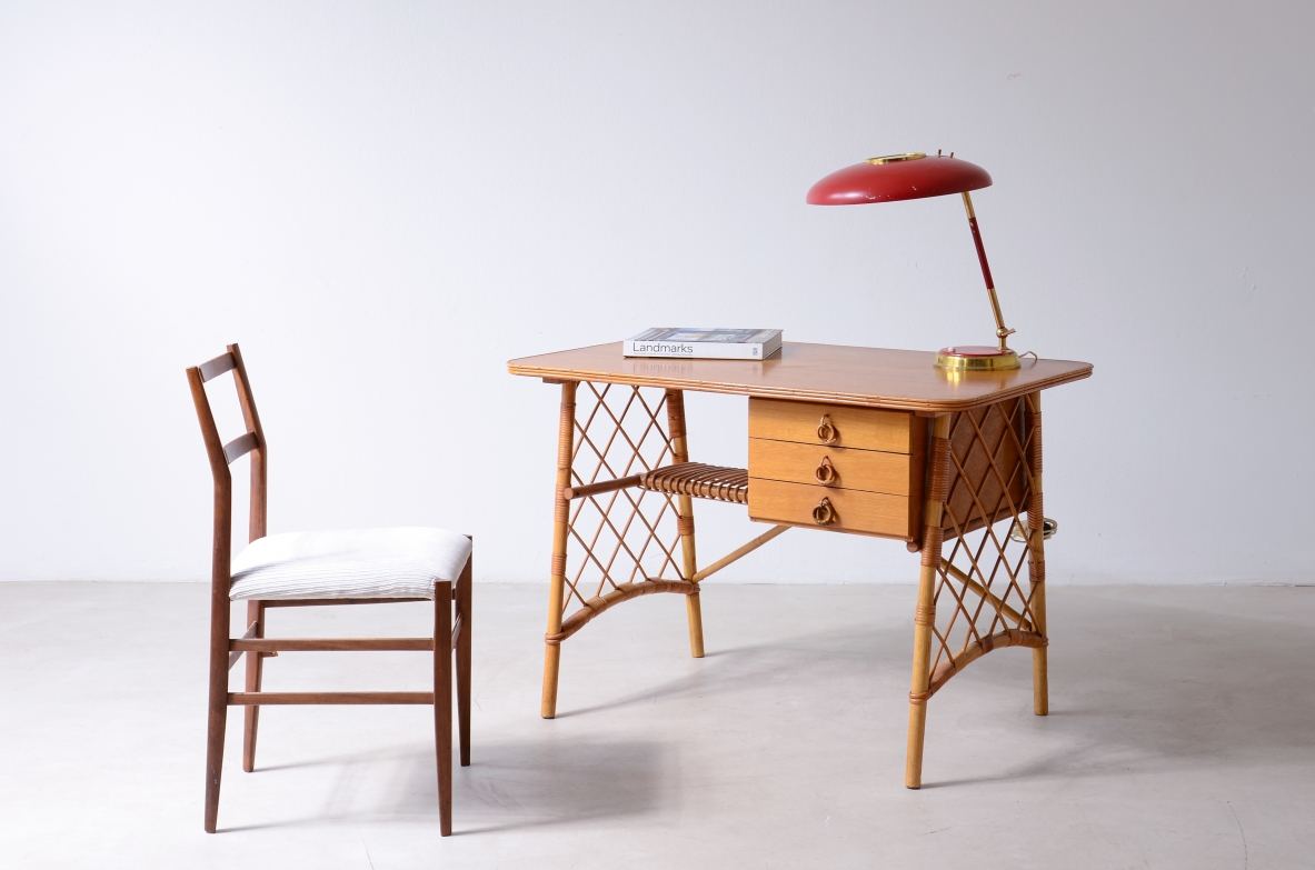 French freestanding writing desk circa 1960 in oak and woven cane, with three side drawers, decorative latticework side supports and rectangular top, photographed in three-quarter view with red lamp.