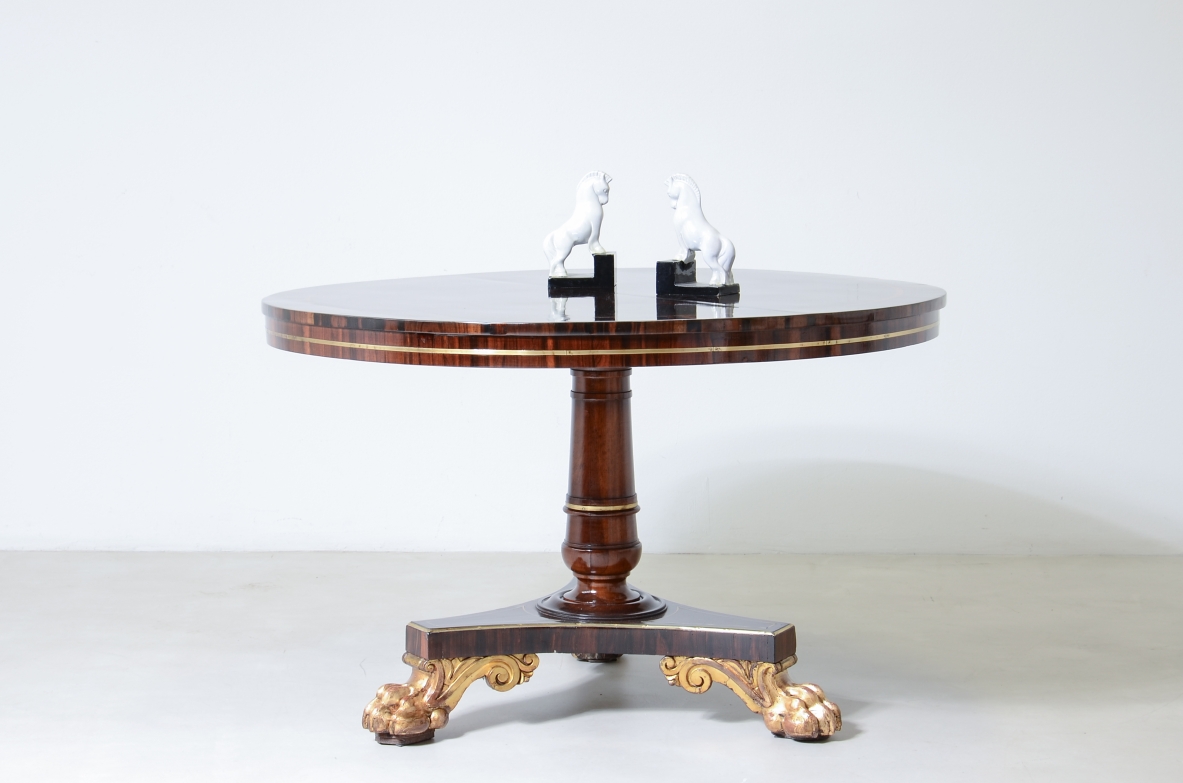 Italian Empire table from Lucca, c. 1820, with a circular exotic wood top edged in brass, a turned central pedestal and a three-legged base ending in large gilded animal paw feet.