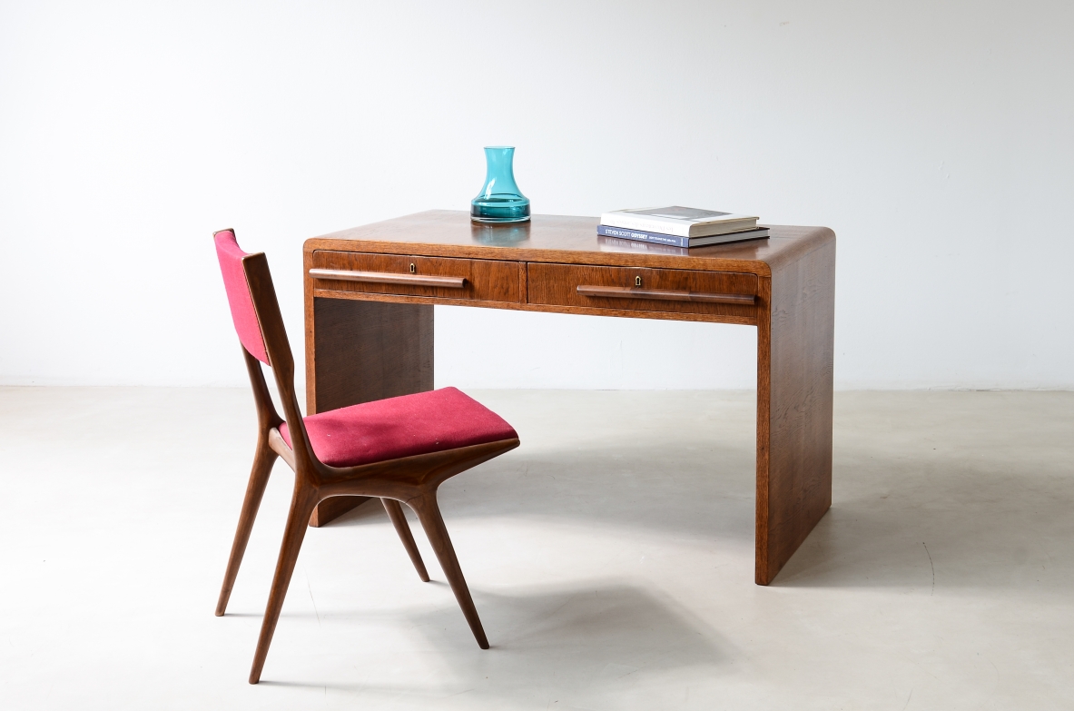 Italian oak writing desk circa 1935 with rectangular top, two drawers and shaped wooden handles, photographed in three-quarter view with red upholstered chair.