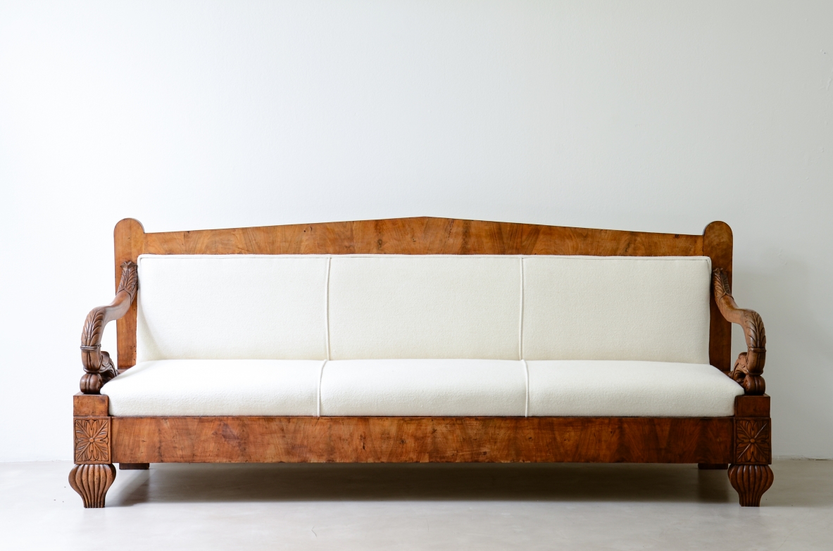 Front view of a Neapolitan Charles X sofa in figured walnut veneer, featuring a neoclassical pediment-shaped back, carved armrests, turned feet and white upholstered seat and backrest.